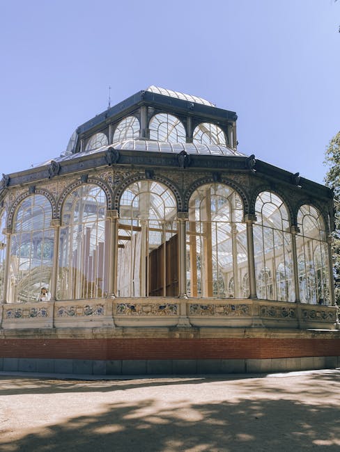 A historic glass greenhouse with ornate metal framing and arched windows, located in a garden setting. The structure features multiple tiers, with the lower level showcasing large, curved glass panels
