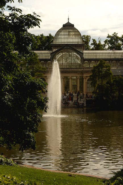 A historic glass greenhouse with ornate metal framing and arched windows, located in a garden setting. The structure features multiple tiers, with the lower level showcasing large, curved glass panels