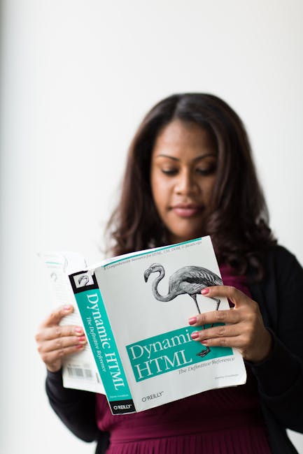 A woman with dark brown hair, wearing a black blazer over a dark pink top, is holding a book titled 'Dynamic HTML' by O'Reilly, featuring a detailed illustration of a flamingo on the cover. She is loo